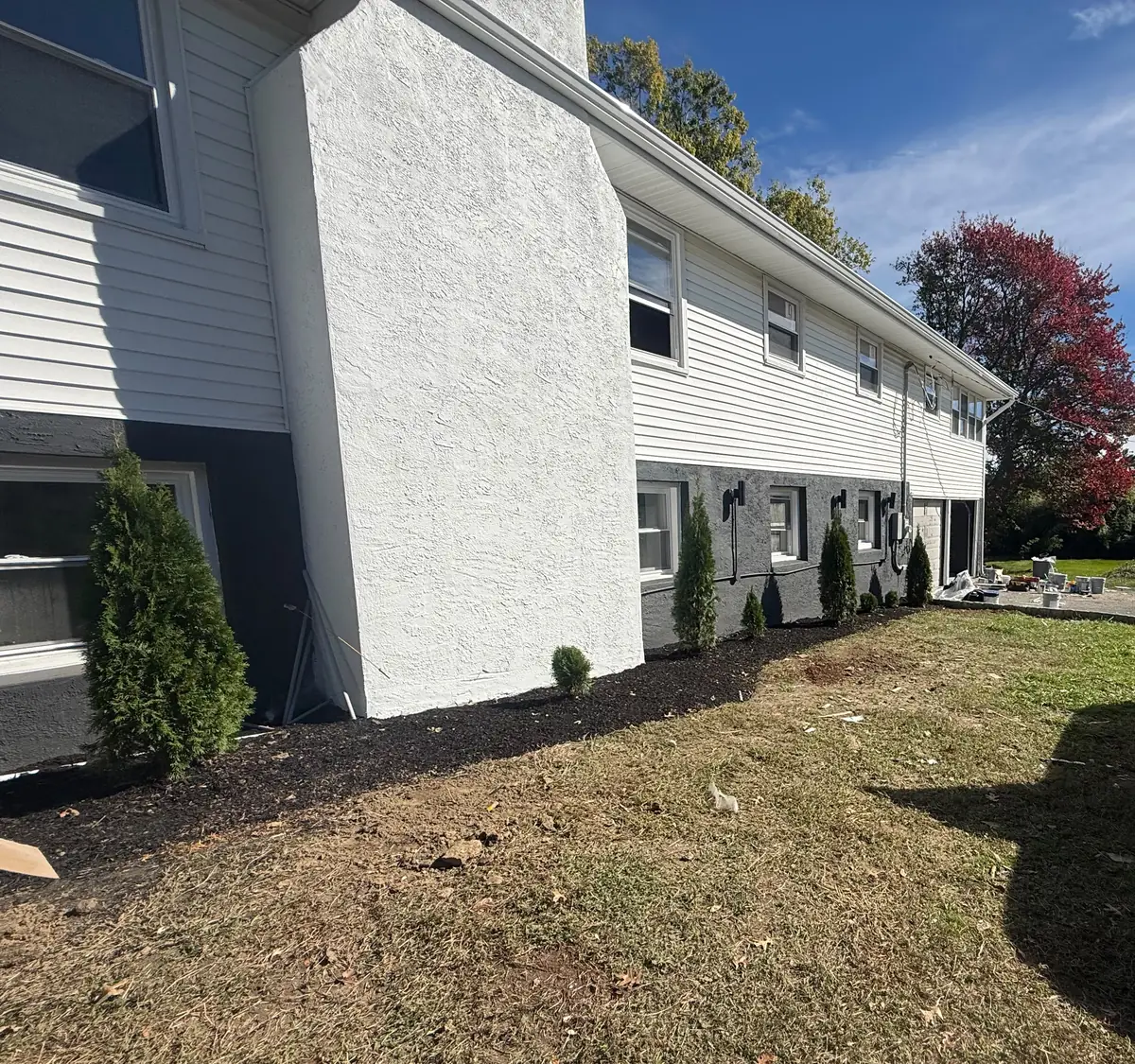 The long side of the house in October 2026, with new siding, new wall lights, fresh mulch beds and evergreen plantings along a wide lawn.