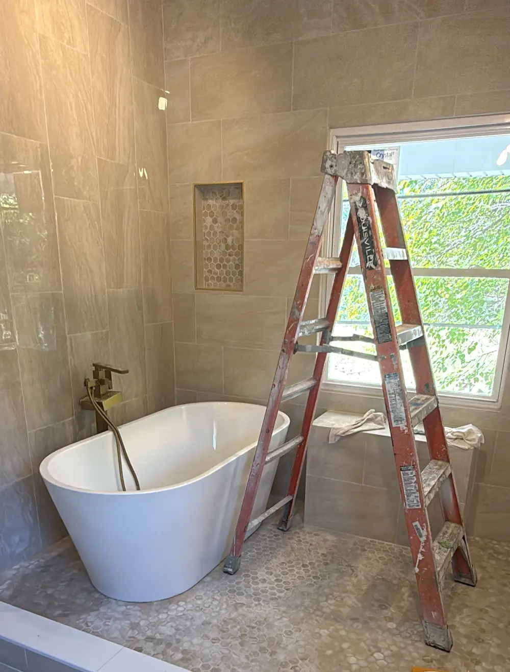 The primary bath wet room with a white freestanding tub, a brushed-gold floor-mounted filler, a hex-mosaic floor and large-format tile walls.